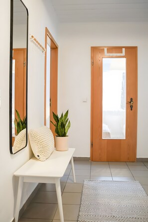 Un couloir avec une porte en bois, un banc blanc avec des coussins, une plante en pot, un miroir et un tapis à motifs.