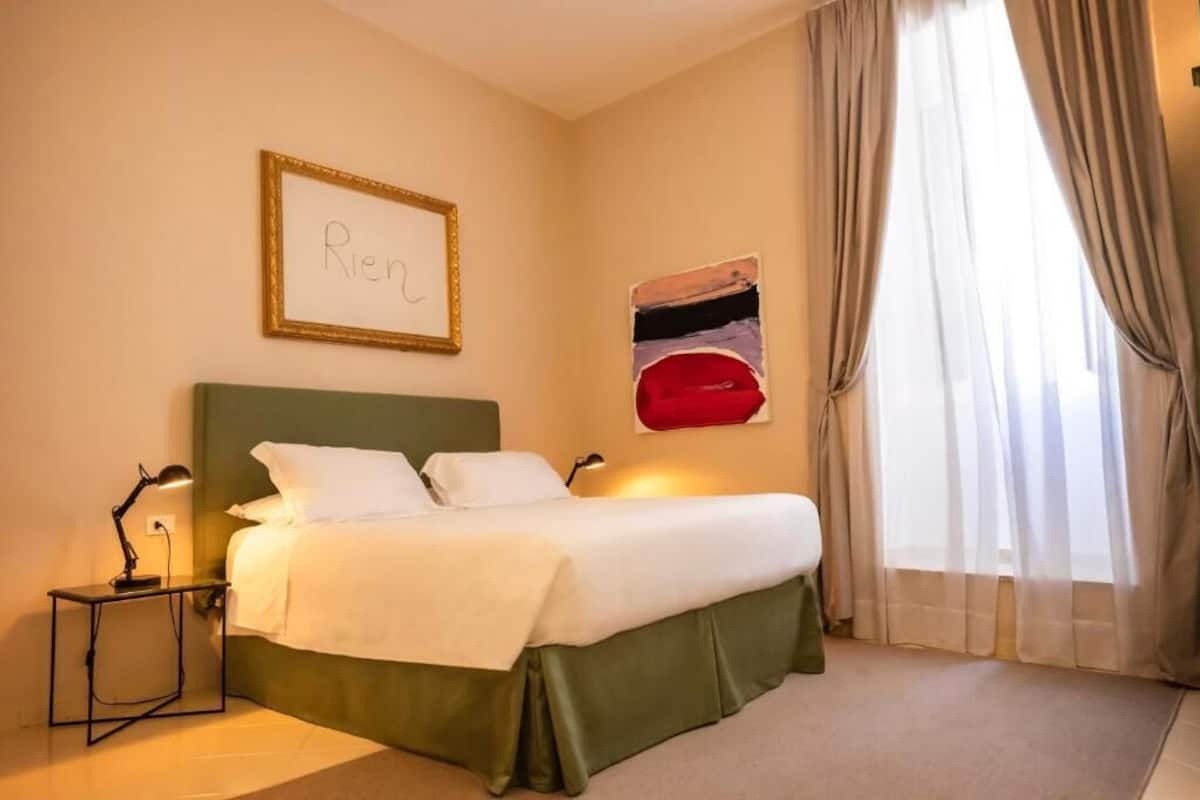 Habitación de hotel con cama, mesita de noche, lámpara, un cuadro en la pared y ventana con cortinas.