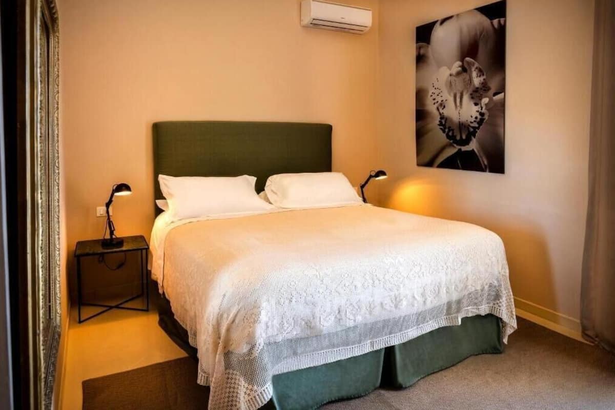Una cama bien hecha con cabecera verde, ropa de cama blanca y cubre lecho de encaje. Hay dos mesitas de noche con lámparas y un equipo de aire acondicionado sobre la cama.