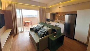 Une chambre d’hôtel moderne équipée d’un canapé, d’une petite table à manger, d’un coin cuisine avec un réfrigérateur et d’une porte-fenêtre coulissante donnant sur un balcon.