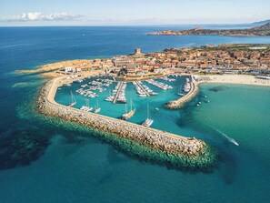 Vista aérea de un pueblo costero con puerto deportivo, espigón rocoso y edificios a lo largo de la costa.