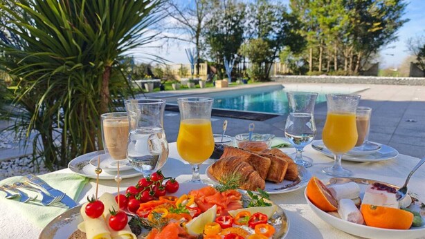 Mesa al aire libre con diversos alimentos, entre ellos salmón ahumado, verduras y pan, junto con vasos de zumo de naranja y agua.