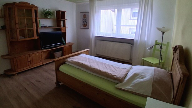 A room with a wooden bed, a television on a wooden stand, a radiator, and a window with curtains.