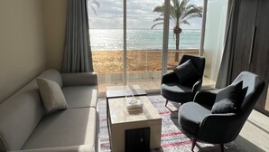 A modern living room with a sofa, armchairs, and a coffee table. Large windows offer a view of the beach and ocean. - Voya Hotel and Suites (Tyre)