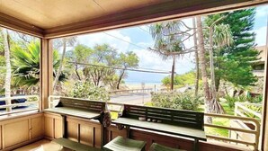 Une pièce avec un banc et deux chaises, un sol carrelé et une vue sur la plage et les arbres, visible à travers de grandes fenêtres.