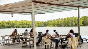 A covered outdoor dining area with tables and chairs, overlooking a body of water and a forested area.