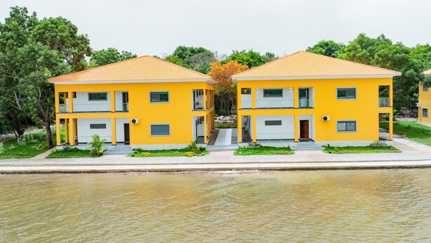 Two yellow buildings with orange roofs, a water body in the foreground, and green trees in the background.