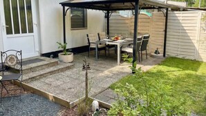 Une terrasse avec une table et des chaises, un espace salon couvert et un petit jardin.
