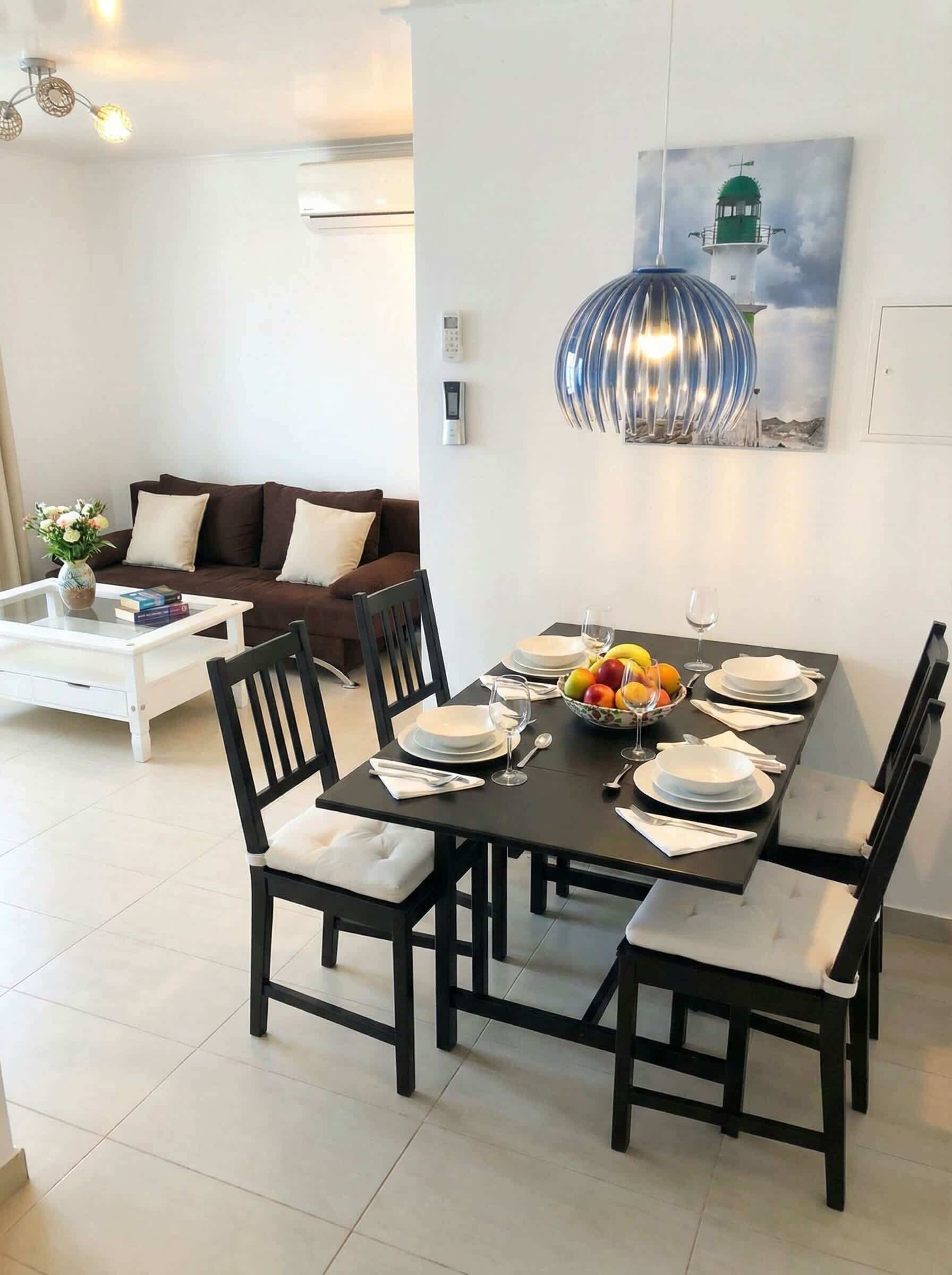 Un coin repas avec une table noire dressée pour six personnes, entourée de chaises noires dotées de coussins blancs. Un tableau représentant un phare est accroché au mur au-dessus de la table.