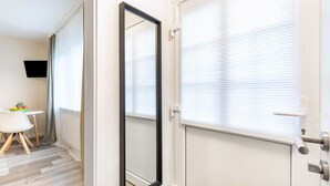 Ein Hotelzimmer mit Holzboden, einer weißen Tür mit Griff, einem großen Spiegel und einem Fenster mit Jalousien.
