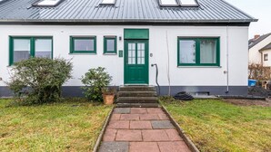 Ein weißes Gebäude mit einer grünen Tür, zwei Fenstern mit grünen Rahmen und einem kleinen Garten mit einem Weg zum Eingang.