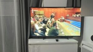 Une pièce avec une télévision posée sur une commode, un lit recouvert d’une couvre-lit à motifs et un rideau.