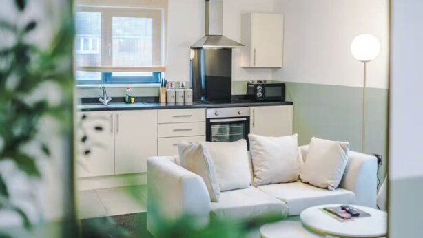 A modern kitchen with white cabinets, a stainless steel range hood, and a microwave. There is a white sofa and a small round table with a magazine. - Business Travellers & Couples with Free Parking (Salford)