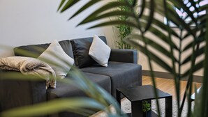 Una sala moderna con un sofá gris oscuro, una pequeña mesita negra y una planta verde grande en primer plano.