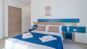 A hotel room with a bed, a blue headboard, a wooden door, a wall-mounted artwork, and a nightstand with a lamp. - Giualè Portopalo '4' with Sea View, Wi-Fi and Air Conditioning (Portopalo di Capo Passero)