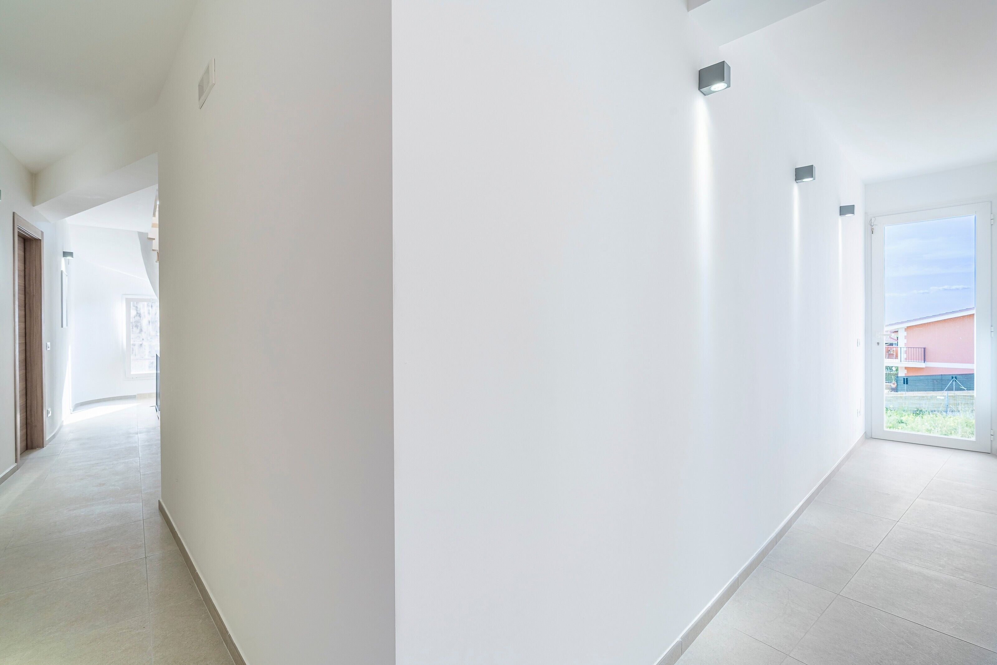 A corridor with white walls and tiled flooring, leading to a room with a large window and a door.