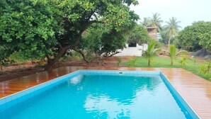 A rectangular swimming pool with a wooden deck surrounding it, set in a landscaped area with trees and a building in the background. - sree veda wellness resort (Vanur)