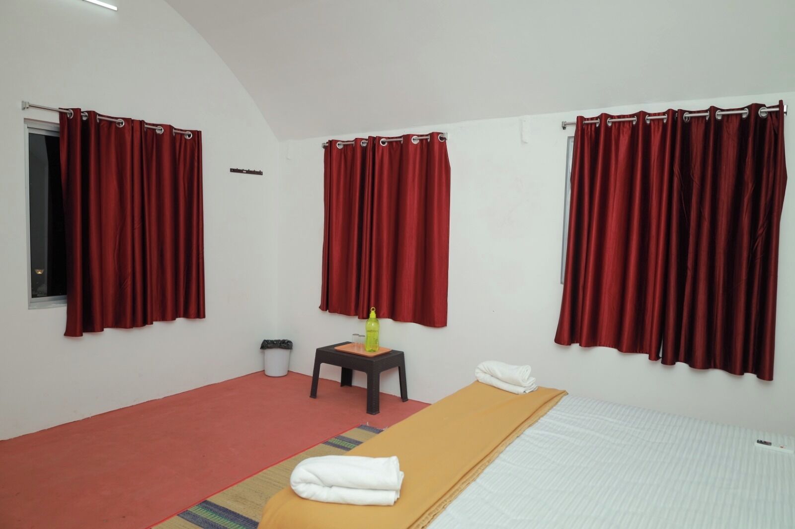 A room with a bed, a small table with a bottle, a trash bin, and red curtains.