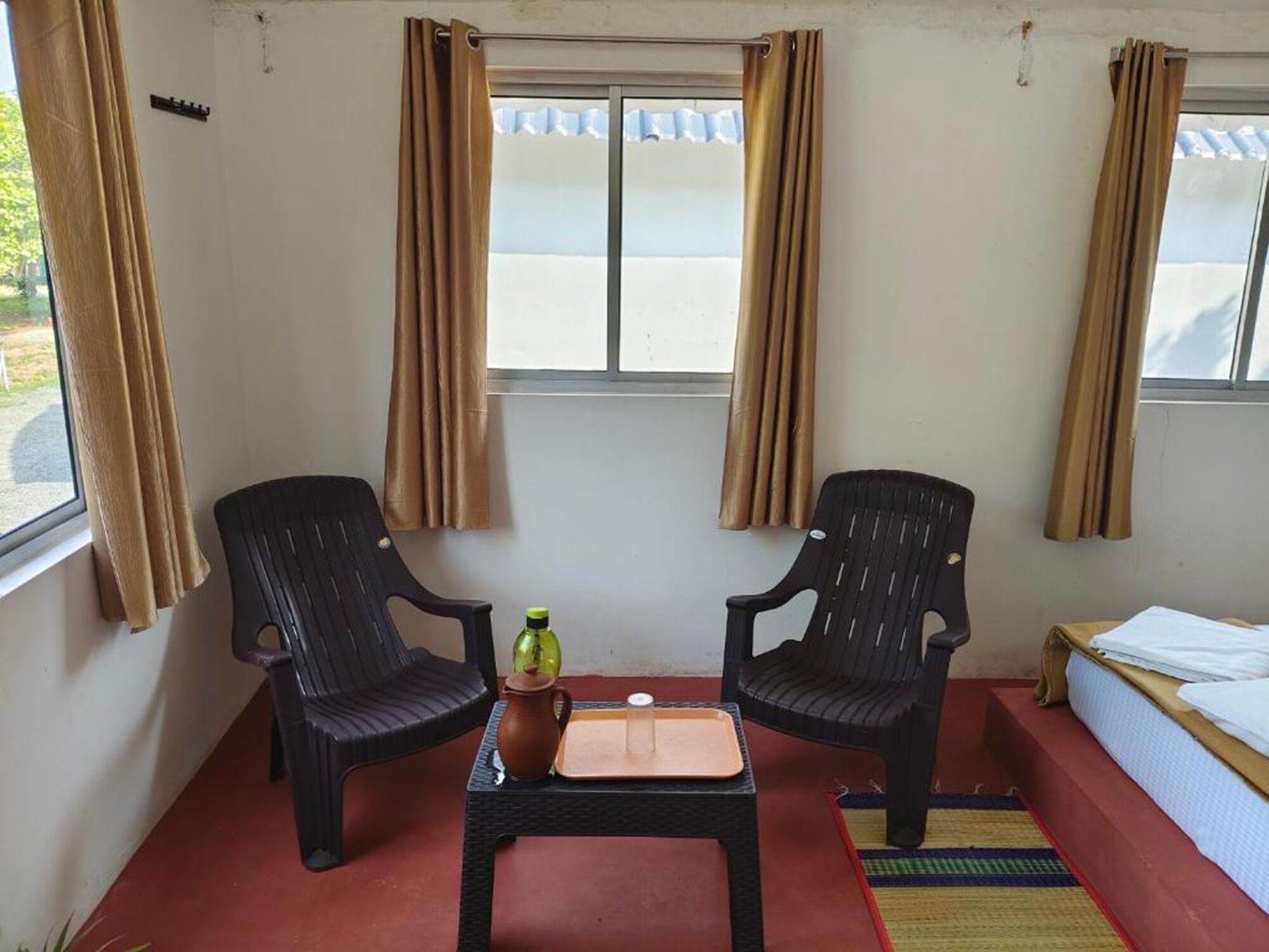 Une petite pièce avec deux chaises noires, une table sur laquelle se trouvent une bouteille et un plateau, un lit et une fenêtre avec des rideaux.