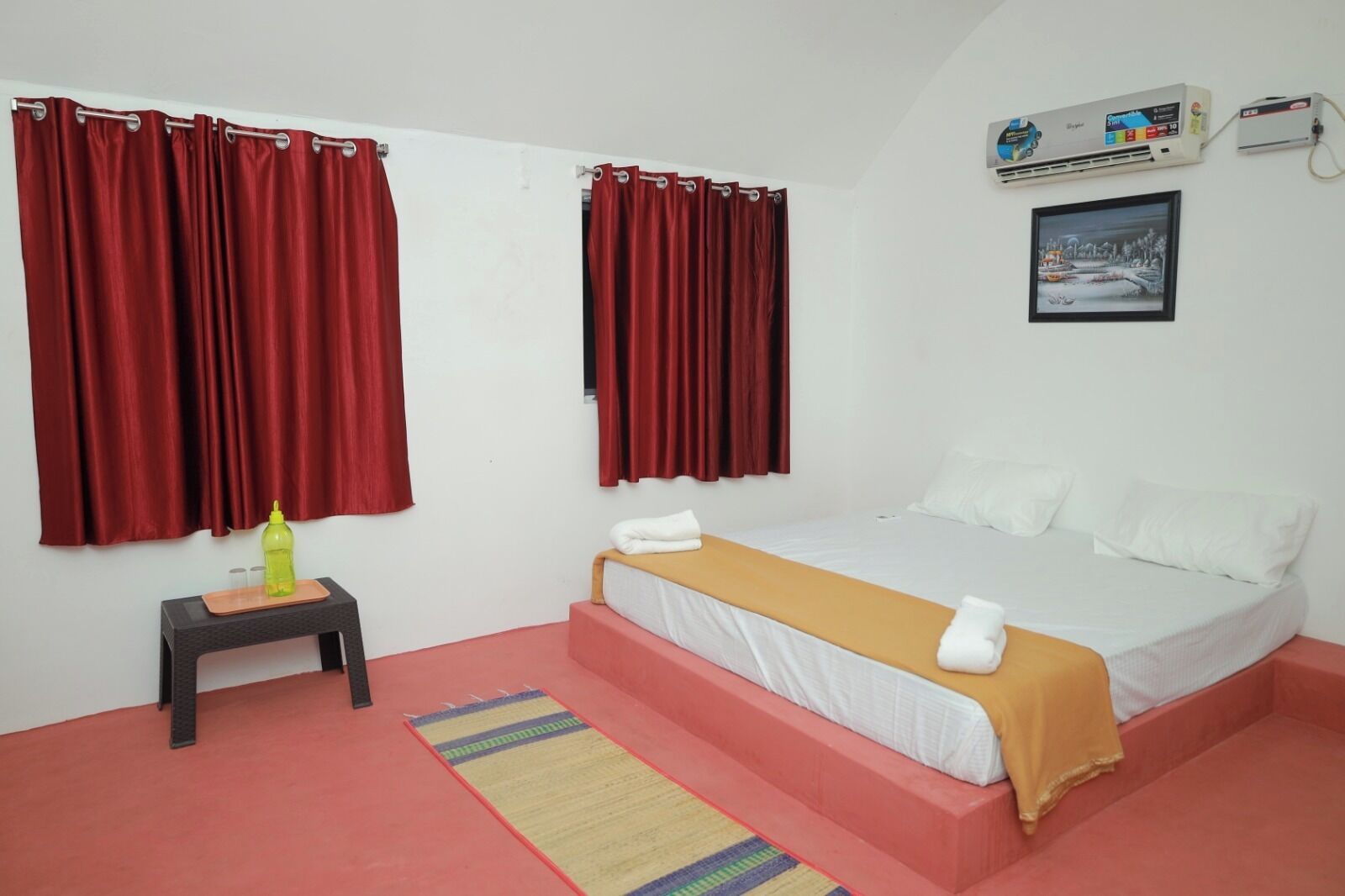 A bedroom with a bed, red curtains, a small table with a bottle, and a picture on the wall.