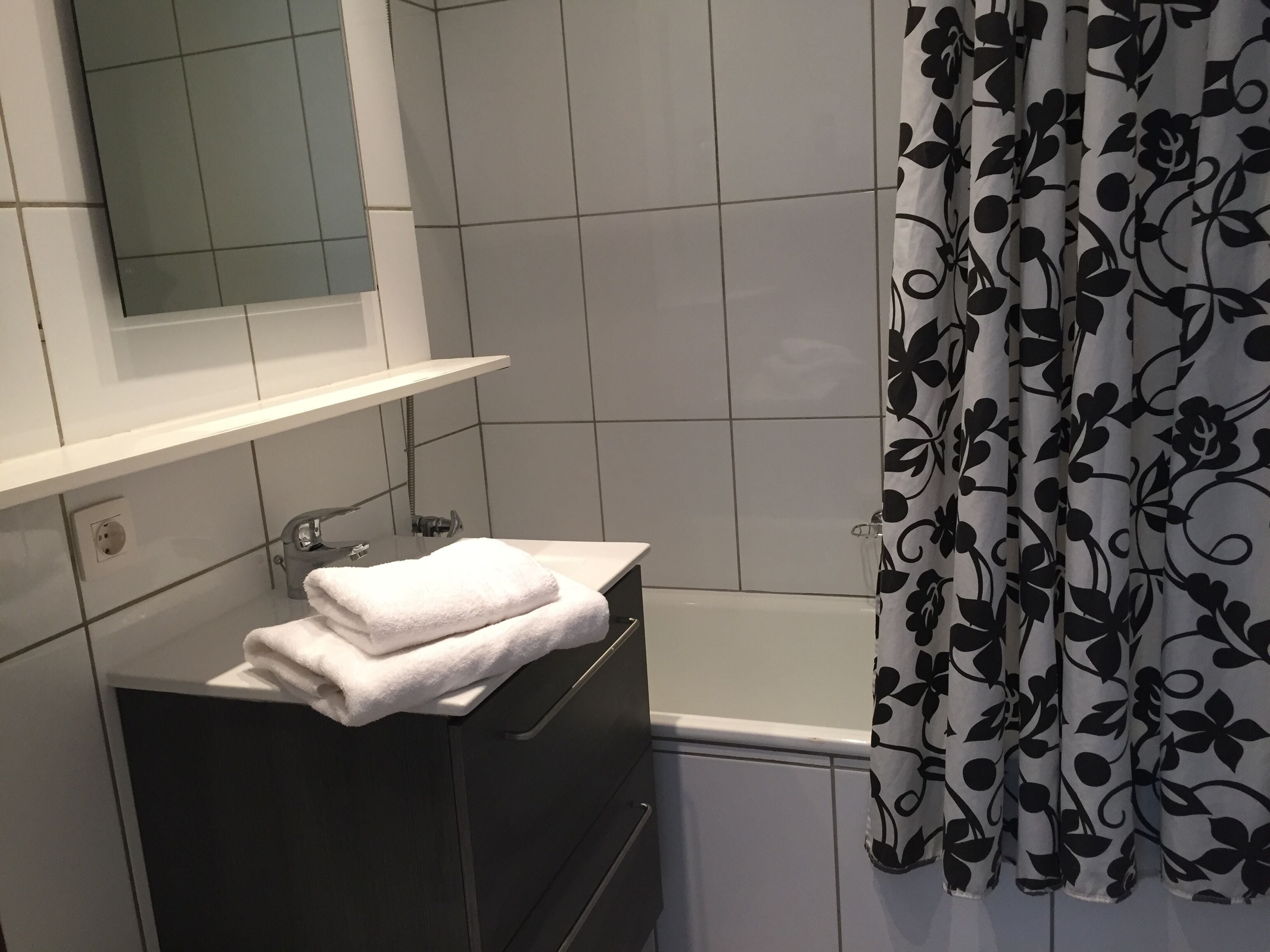 Ett badrum med ett vitt handfat, en spegel ovanför, en handdukshängare med hopvikta handdukar och en duschdraperi med ett svart blommönster.
