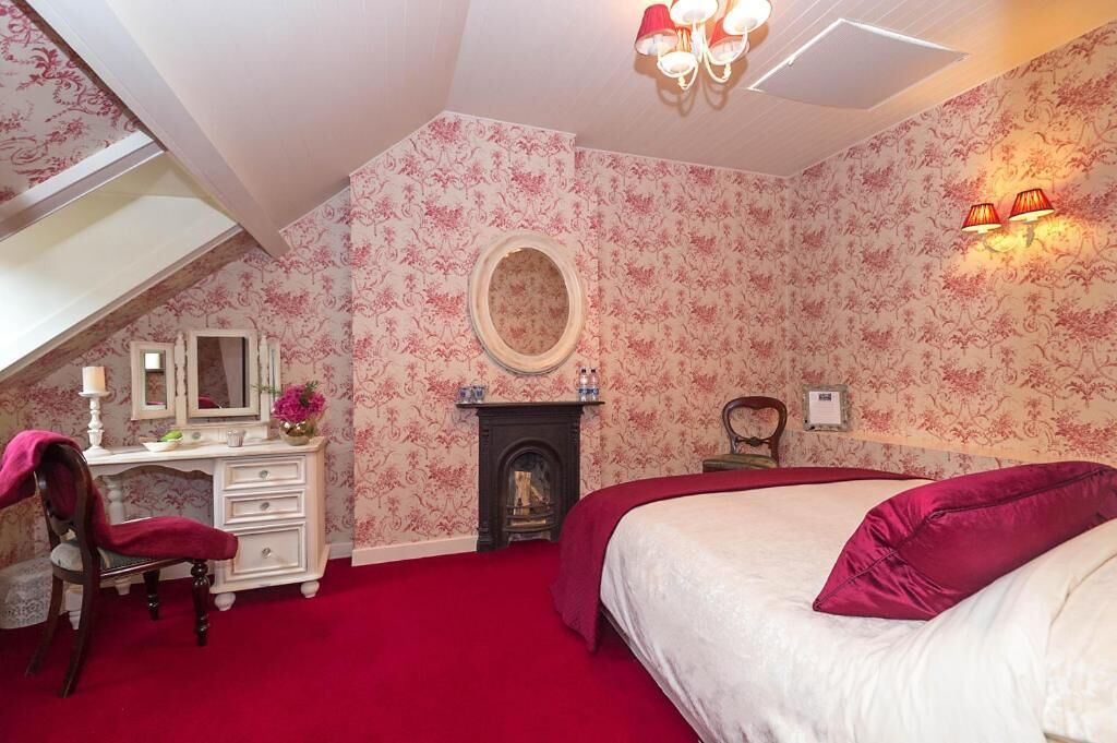 A bedroom with a red carpet, a bed with a red pillow, a dresser, a mirror, a fireplace, and a chandelier.