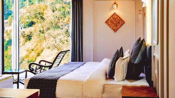 A bedroom with a bed, a chair, a table, and a view of a mountainous landscape. - Sunoraa Luxury Hotel And Spa (Mussoorie)