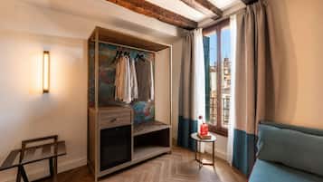 A room with a wooden wardrobe, a small table with a vase, a sofa, and a view of buildings through a window.