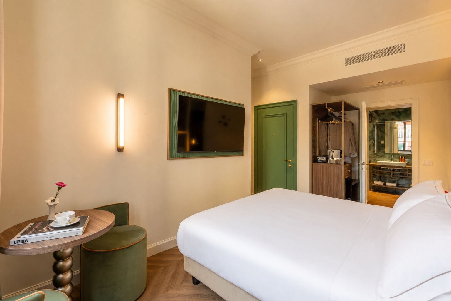A hotel room with a bed, a desk with a chair, a green door, and a bathroom visible through an open door.