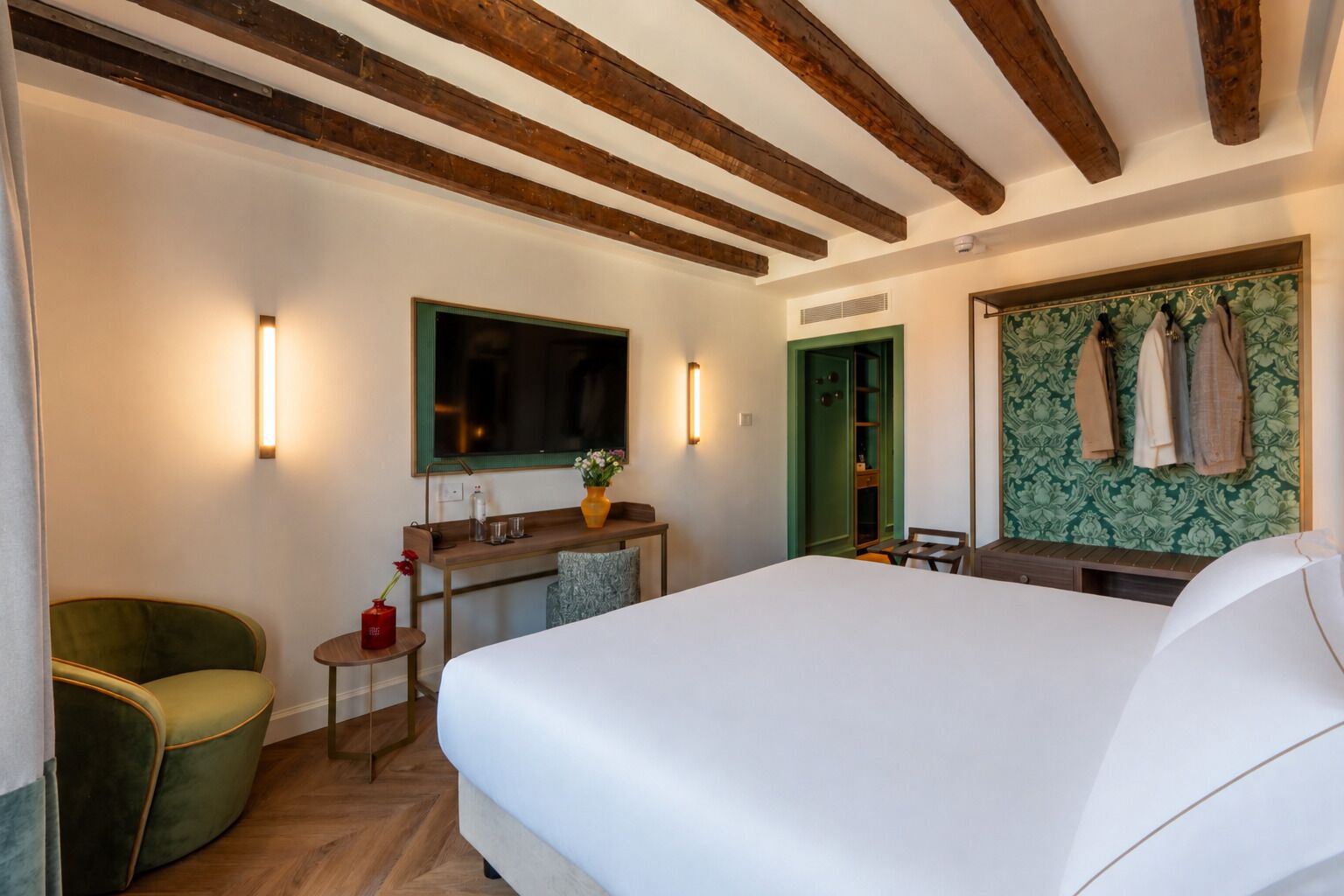 A hotel room with a large bed, a desk with a chair, a mirror, and a wardrobe with hanging clothes.
