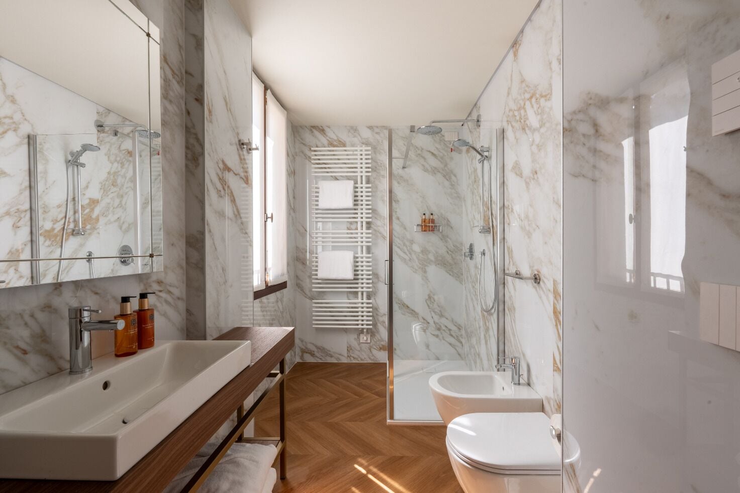 Un bany modern amb una paret de marbre, un moble de tocador de fusta, una cabina de banyera de vidre i un vàter.