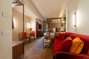 A hotel room with a red sofa, a dining area with a table and chairs, a kitchen area, and a hallway leading to another room.