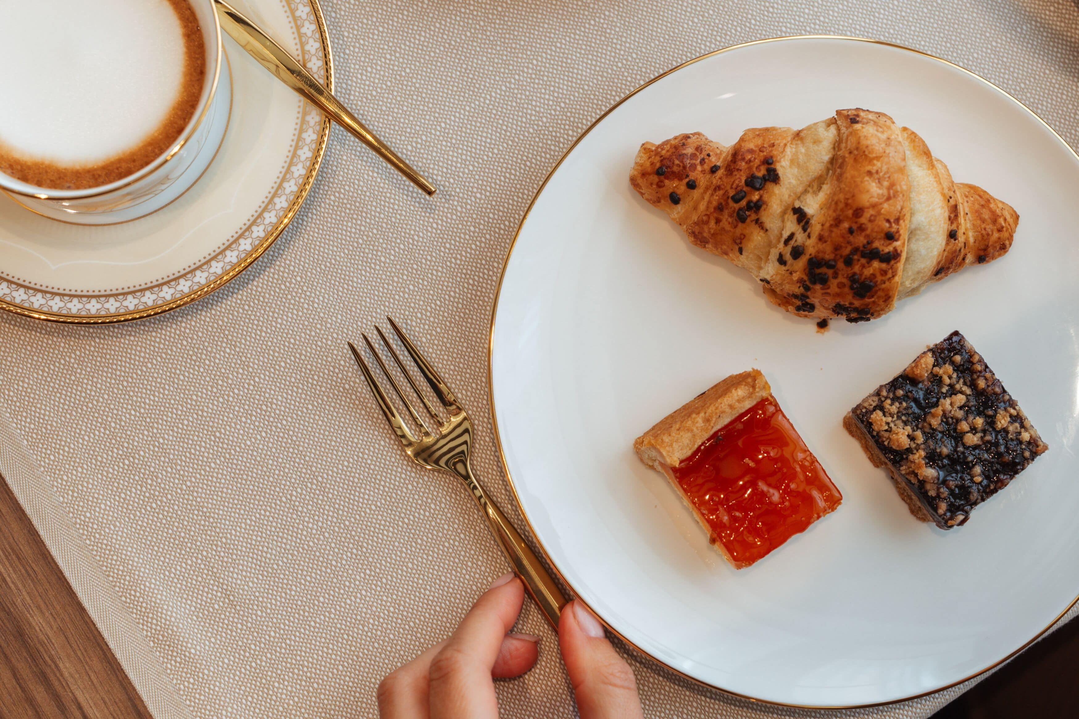A plate with a croissant, a tart, and a dessert on a table with a cup and a gold fork.