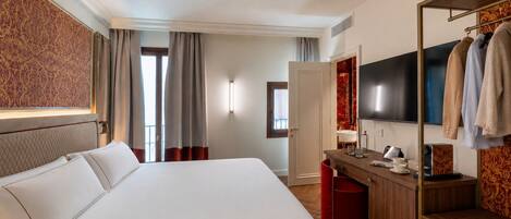 A hotel room with a large bed, a desk with a flat-screen TV, a mirror, and a wardrobe with hanging clothes.