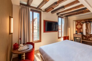 A hotel room with a bed, a table with a newspaper, a red chair, a TV on the wall, and a window with curtains. - Hotel San Samuele (Venice)