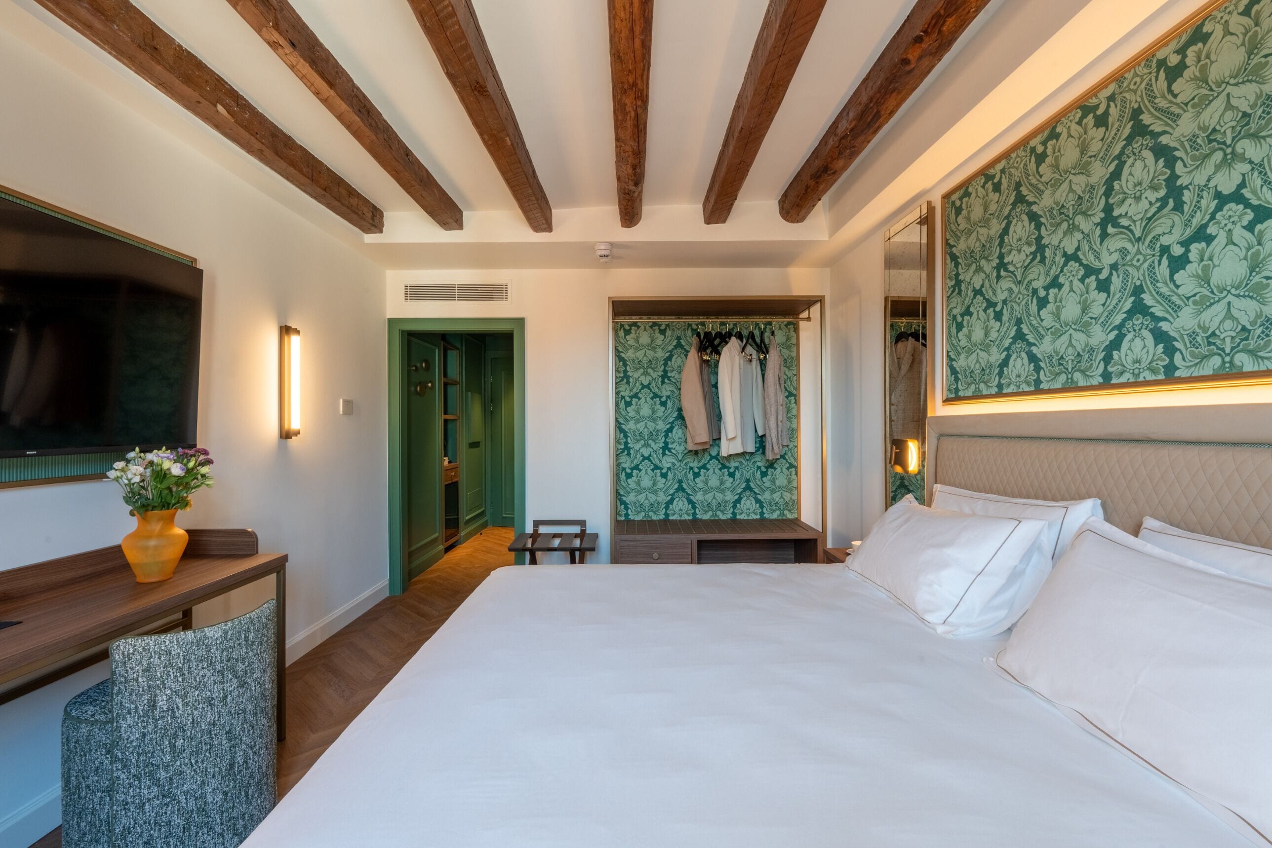 A hotel room with a large bed, a vanity with a mirror, a green patterned wall, and wooden ceiling beams.