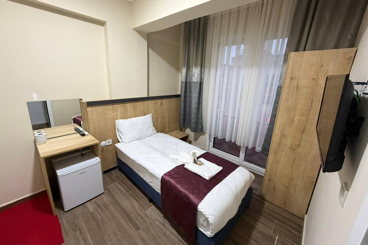 Ein Hotelzimmer mit einem Einzelbett, einem Schreibtisch mit Stuhl, einem kleinen Kühlschrank und einem großen Fenster mit Vorhängen.