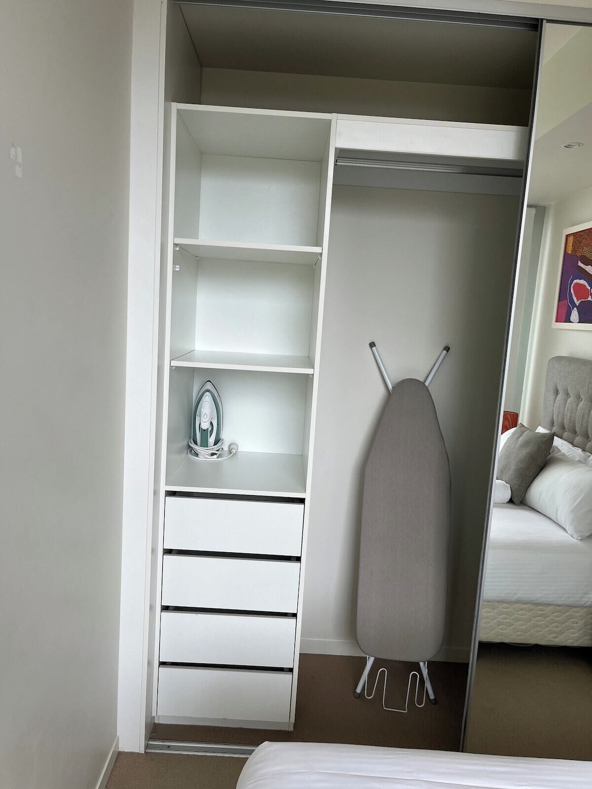 A bedroom with a bed, a wardrobe with open doors, and a hanging ironing board.