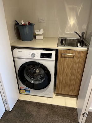 A compact laundry area with a front-loading washing machine, a sink, and a cabinet. - Tasha's Apartments on Morphett (Adelaide)