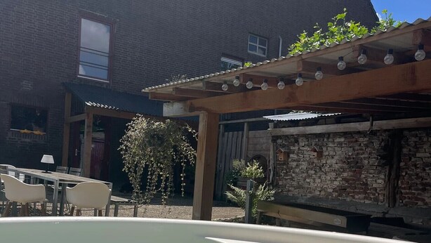 Uma área de piscina com pérgola, um edifício de tijolos ao fundo e um pequeno jardim com plantas.