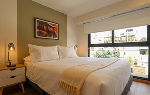 A bedroom with a large bed, a nightstand with a lamp, a painting on the wall, and a view of buildings through the window. - w Tranquil 2BR w Balcony in Miraflores (Lima)