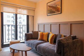Ein modernes Wohnzimmer mit einem grauen Sofa, einem runden Holztisch und einem großen Fenster mit Blick auf ein Gebäude.