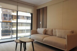 Een moderne woonkamer met een bank, een klein rond tafeltje en een groot raam met uitzicht op een gebouw.