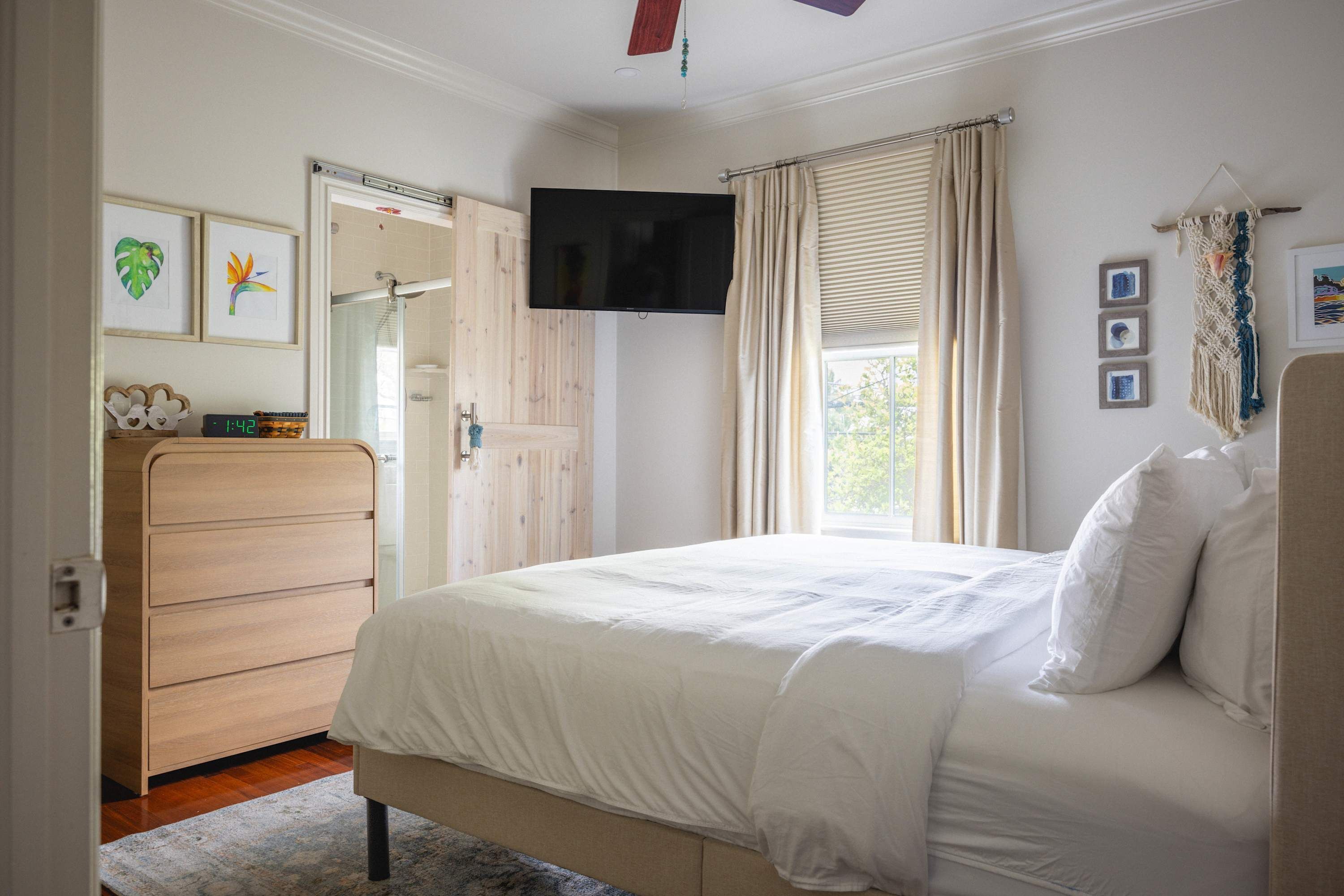 A bedroom with a bed, a TV mounted on the wall, a wooden dresser, and a window with curtains.