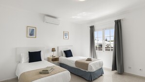 Une chambre d’hôtel avec deux lits, une fenêtre avec des rideaux, un appareil de climatisation et des tableaux encadrés au mur.
