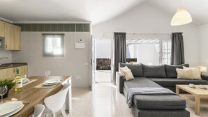 Un salon moderne avec un canapé d’angle gris, une table à manger en bois avec des chaises, un coin cuisine et une grande fenêtre.
