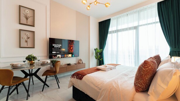A modern hotel room with a large bed, a dining table with chairs, a TV mounted on the wall, and a large window with curtains. - Luxury Studio with Pool - Azizi Vista (Dubai)