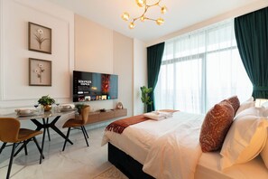Et moderne hotelværelse med en stor seng, et spisebord med stole, et TV ophængt på væggen og et stort vindue med gardiner.