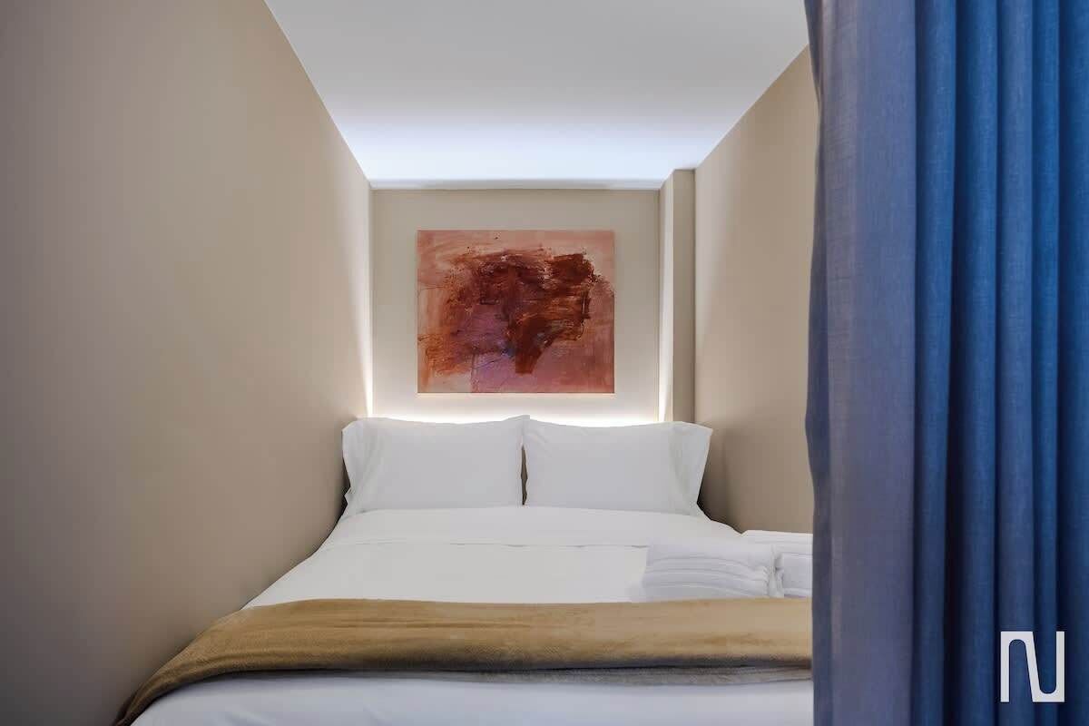 A hotel room with a bed, white linens, a brown throw blanket, a ceiling light, and a wall-mounted abstract painting.