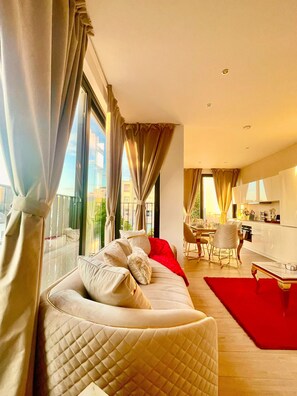 A modern living room with a large window, a sofa with cushions, a dining area with a table and chairs, and a kitchen area. - One Lux Stay Near Antwerp Central - 2 Bedroom 1.5 Bathroom (Antwerp)